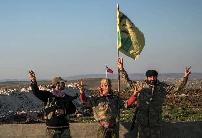 In this Sunday, Feb. 22, 2015, file photo, Syrian Kurdish militia members of YPG make a V-sign next to poster of Abdullah Ocalan, jailed Kurdish rebel leader, and a Turkish army tank in the background in Esme village in Aleppo province, Syria. 