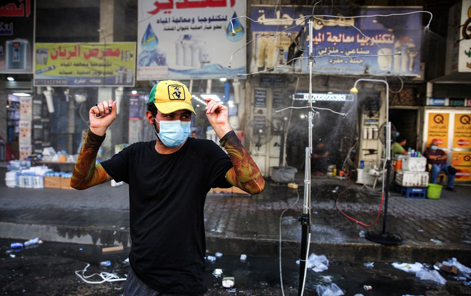 A man clad in mask due to the COVID-19 coronavirus pandemic stands next to sprinklers along Sinak street in Iraq's capital Baghdad on August 9, 2020, to cool off due to extremely high temperature rises amidst a heatwave. Ahmad al-Rubaye / AFP
