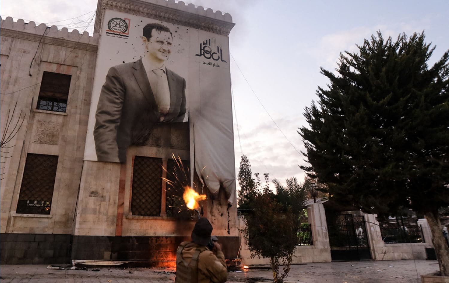 چەکدارێک دوای رووخانی، تەقە لە وێنەیەکی بەشار ئەسەد دەکات کە لەسەر باڵەخانەیەک کێشراوە. وێنە: AFP