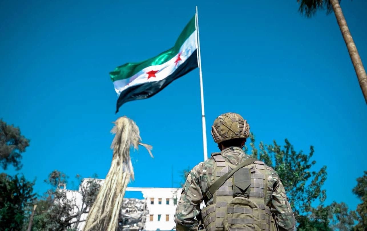 سەربازێکی سووریا لە بەردەم ئاڵای وڵاتەکە. 