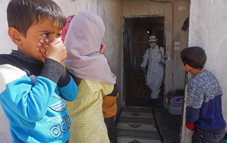 Syrian children watch a member of the Syrian civil defence disinfecting a former school building currently inhabited by displaced families in Binnish, Idlib, March 26, 2020, Photo: Muhammad Haj Kadour / AFP