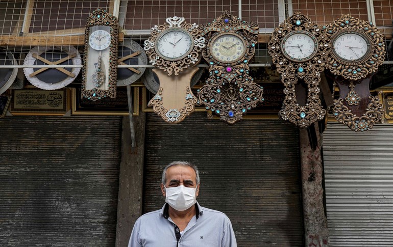 A man wearing a face mask poses for a picture beneath hanging clocks outside a closed shop in the Kurdistan Region capital of Erbil on April 16, 2020. Photo: Safin Hamed / AFP